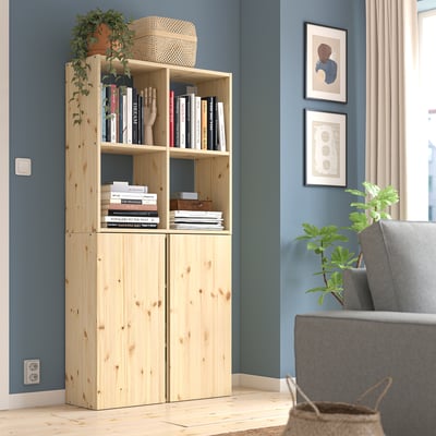 Lichthouten IVAR boekenkast tegen blauwe muur, gevuld met boeken. Naast een grijze stoel, kleine plant en ingelijste foto's.
