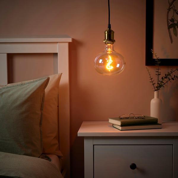 Slaapkamer met warme JÄLLBY lamp boven nachtkastje met boek en vaas, bed met twee kussens, perzik muren.