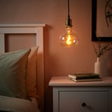 Slaapkamer met warme JÄLLBY lamp boven nachtkastje met boek en vaas, bed met twee kussens, perzik muren.