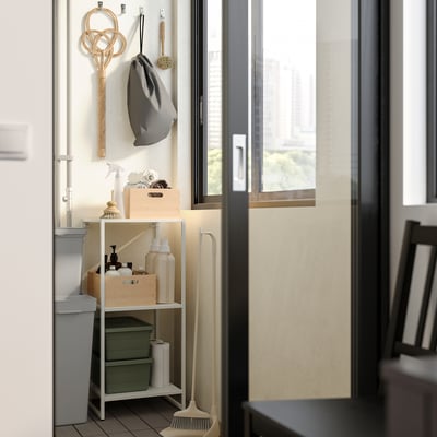 Klein opbergrek in de badkamer met planken met schoonmaakspullen, naast een raam met een dweil en haken voor accessoires, alles netjes georganiseerd.