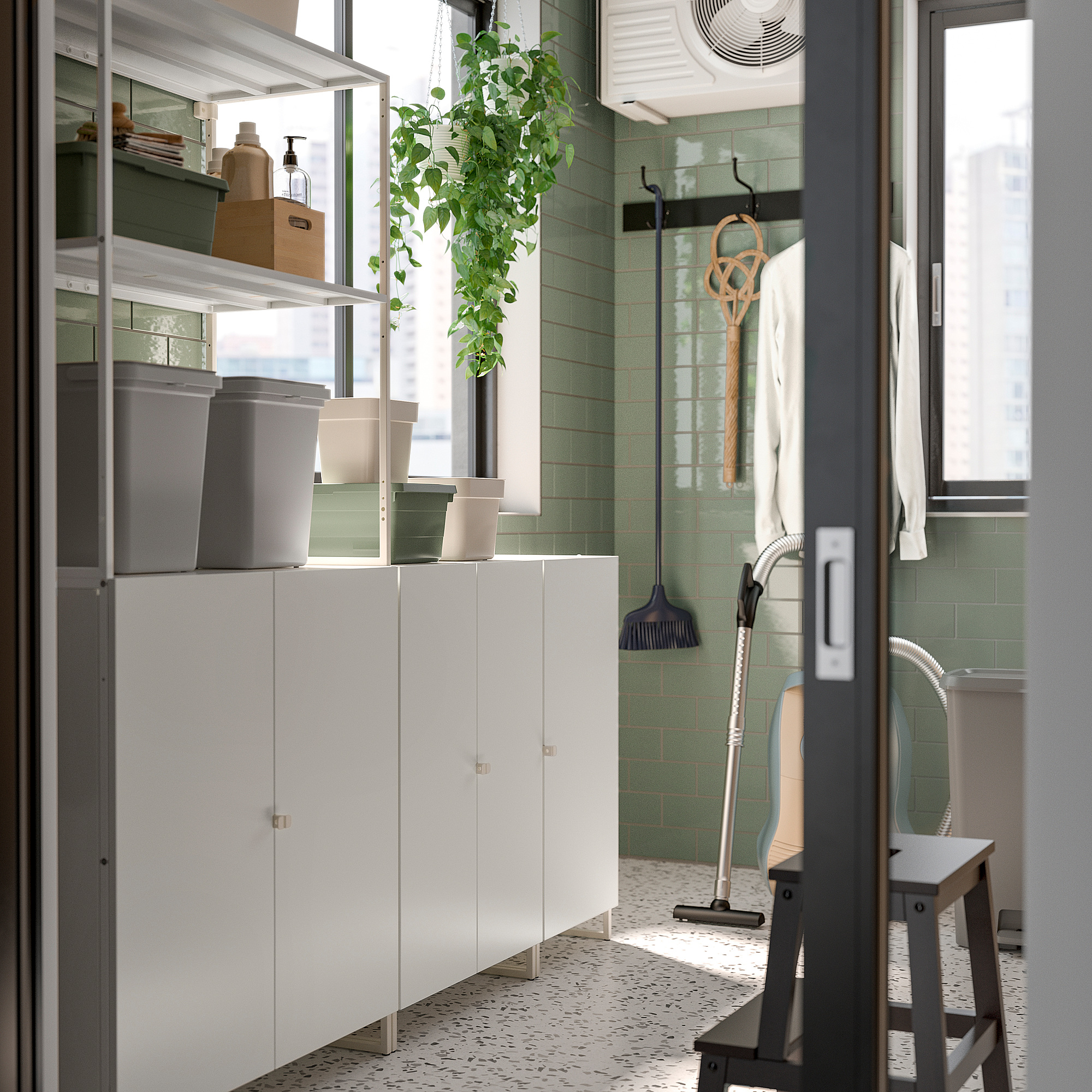 Moderne bijkeuken met JOSTEIN witte opbergkasten, grijze bakken, groene tegels, hangplanten en schoonmaakgereedschap.