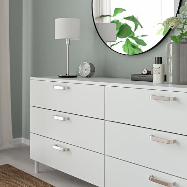 Wit dressoir met zes lades, zilveren moderne handgrepen. Bekroond met een lamp, klok en parfum, weerspiegeld een plant in een ronde spiegel.