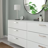 Wit dressoir met zes lades, zilveren moderne handgrepen. Bekroond met een lamp, klok en parfum, weerspiegeld een plant in een ronde spiegel.