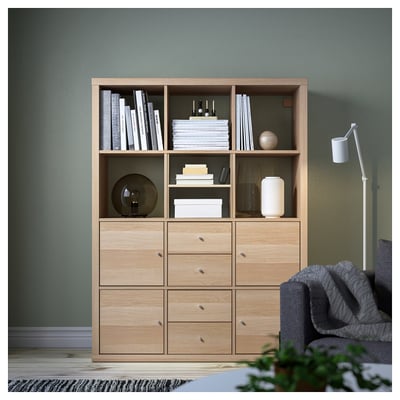 KALLAX lichte houten boekenplank met planken en lades, staande tegen groene muur, omgeven door een grotere grijze stoel en kleine plant.