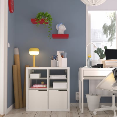 Thuiskantoor met KALLAX-plank, boeken, heldere lamp, rode plantenplanken, klok, bureau, computer. Blauwe muur, houten vloer.