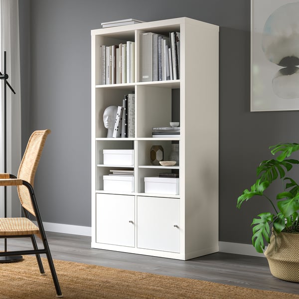 Witte KALLAX boekenplank met boeken, decor en opslag in de buurt van grijze muur. Rieten stoel en plant in de buurt.