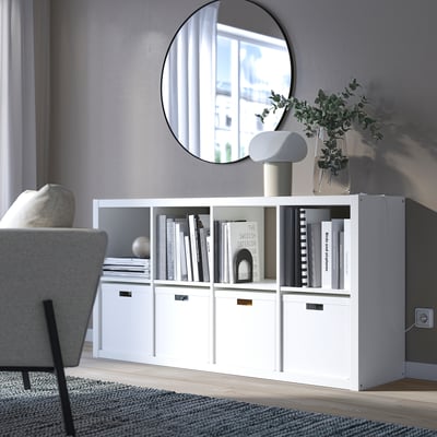 Witte ikea KALLAX plank met boeken, decor, plant en lamp. Grote spiegel reflecteert raam. Grijze bank, vloerkleed, gezellige minimalistische ruimte.