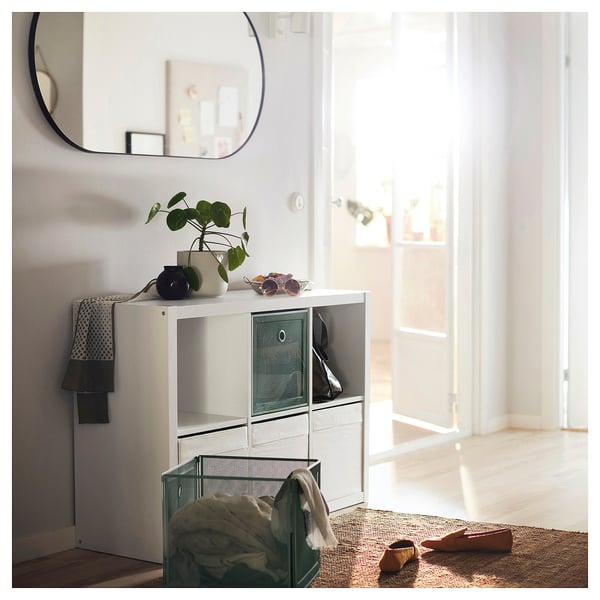 Helder verlichte entree met KALLAX witte rekken, groene mand met kleding, potplant, zonnebril, spiegel boven, zwarte tas, jute mat, schoenen.