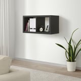 Een KALLAX zwarte wandplank met boeken, een klok en een ingelijste foto. Een crèmekleurige bank en een potplant staan eronder.