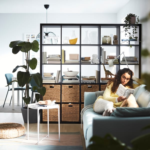 De vrouw leest boek op blauwe bank dichtbij zwarte KALLAX plank met rieten manden, installaties, en decor.