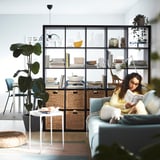 De vrouw leest boek op blauwe bank dichtbij zwarte KALLAX plank met rieten manden, installaties, en decor.