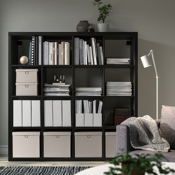Zwarte KALLAX boekenkast met planken, stoel en lamp.