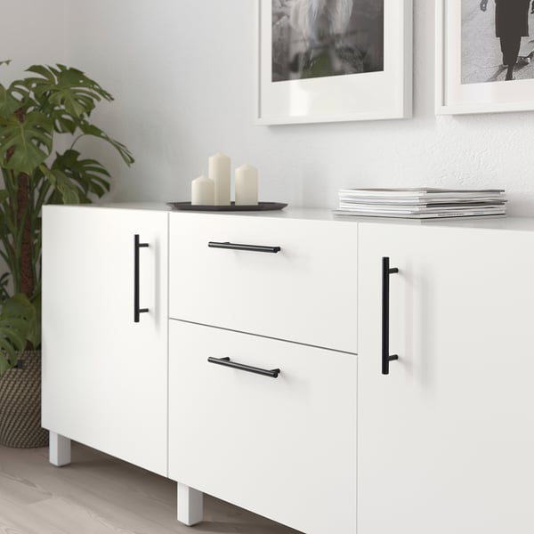 Moderne witte kast met zwarte handgrepen, met kaarsen en boeken, naast een grote lommerrijke plant.