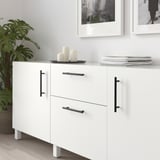 Moderne witte kast met zwarte handgrepen, met kaarsen en boeken, naast een grote lommerrijke plant.