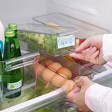 Persoon organiseert koelkast met duidelijke, stapelbare opbergdozen. Etiketten op dozen voor gemakkelijke identificatie.