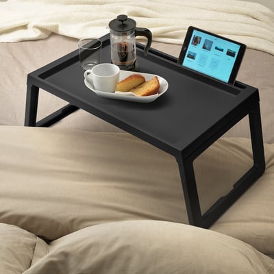 Klein zwart dienblad voor koffie, cake en tablet op een gezellig bed met beige kussens.