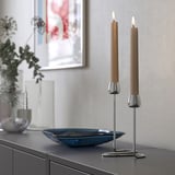 KLOKHET kaarsen, zilveren houder, twee beige kaarsen op moderne kast, kleine schaal, decoratieve vaas links, kunst aan de muur, gezellige woonkamer opstelling.