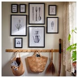 Zwart-ingelijste kunst met bestek & bloemmotieven. Houten peg rek met manden, bezem, kanten gordijn en plant.