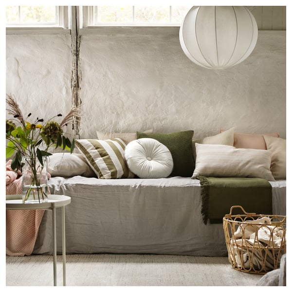 Lichte woonkamer heeft een wit KRANSBORRE rond kussen met groene accenten op een neutrale bank, naast een bijzettafel met bloemen en een gevlochten mand.