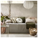 Lichte woonkamer heeft een wit KRANSBORRE rond kussen met groene accenten op een neutrale bank, naast een bijzettafel met bloemen en een gevlochten mand.