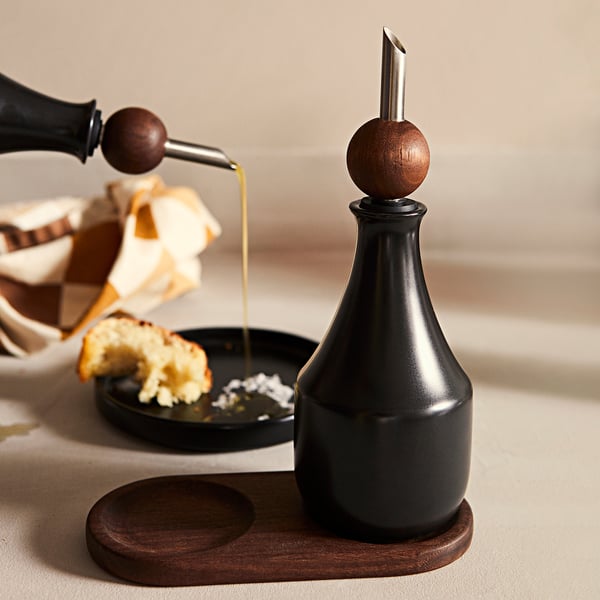 Zwarte KRÖSAMOS oliedispenser met houten stop op dienblad. Eten in kom ernaast.