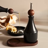 Zwarte KRÖSAMOS oliedispenser met houten stop op dienblad. Eten in kom ernaast.