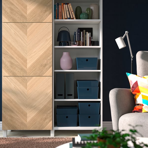 Moderne woonkamer met een blauwe KUGGIS-doos op de plank tussen boeken en decoratie, licht houten paneel, donkere wand, gezellige fauteuil met kleurrijke kussens.