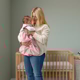 De vrouw die van de blonde een baby in een roze slaapzak voor een houten voederbak houdt.