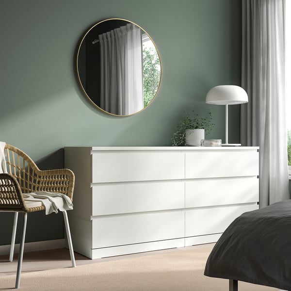 Moderne slaapkamer met wit LINDBYN-dressoir, spiegel met gouden lijst, groenblauwe muur, rieten stoel en weerspiegeld raamgroen.