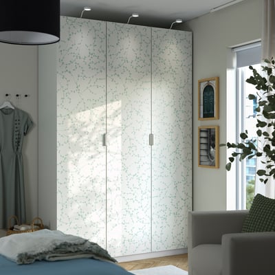 Grote witte LINNABERG kast met groen bladpatroon. Hoog, smal, past tussen bed en grijze bank.