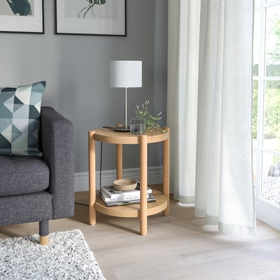 Ronde houten LISTERBY bijzettafel. Lichtbruin, eenvoudig design, naast grijze stoel en witte lamp.