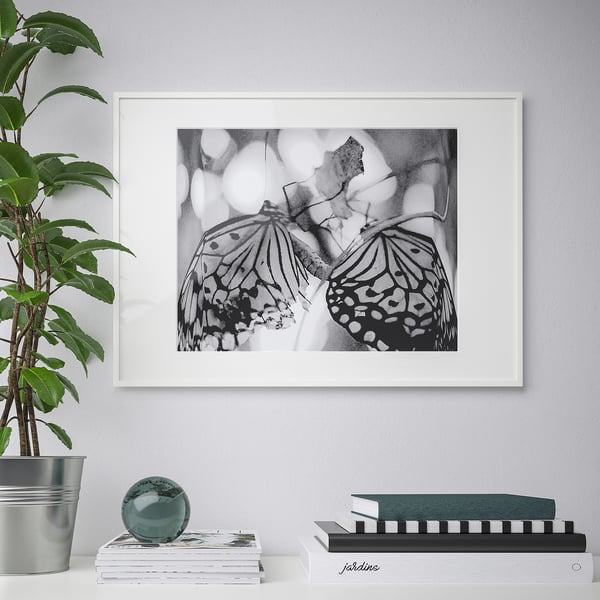 Ingelijste vlinderkunst hangt boven gestapelde boeken en een plant. Het frame is dun en beschermend, geschikt voor verschillende fotoformaten.