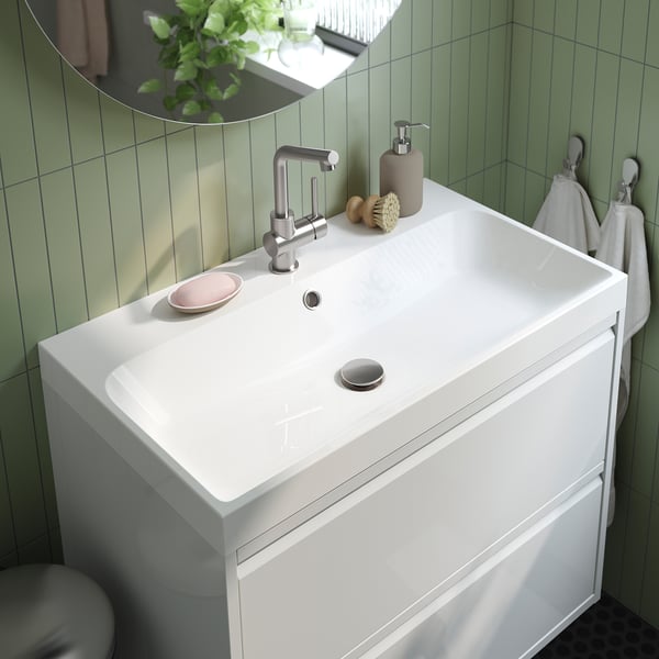 Moderne wastafel in de badkamer met strakke kraan, zeepdispenser en handdoeken. Groen betegelde wanden en witte kast.