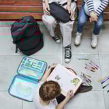 Kinderen op bankje, één tekening met open MÅLA kunstkoffer. Kleurrijke voorraden en kunstwerken op de grond, grote rugzak naast hen.