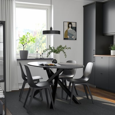 Moderne eetopstelling met ronde tafel, vier zwarte stoelen, hanglamp, grijs vloerkleed en groot raam met natuurlijk licht en kunstwerken.
