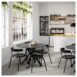 Minimalistisch café met zwarte tafels, grote ramen, krijtmenu en planten. Wit betegelde wanden met koffiestation.