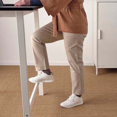 Een persoon die op een hoge tafel leunt met één voet op een ingebouwde voetsteun, waarmee het gebruik ervan voor zowel zittend als staand werk wordt gedemonstreerd.