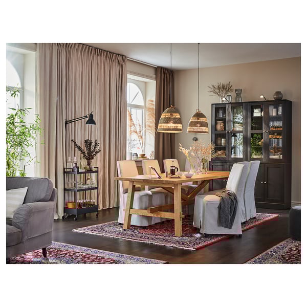 Rustieke eetkamer met houten tafel, grijze stoelen, zwarte kast en houten hangers. Roze gordijnen, kleurrijk tapijt en planten voegen warmte toe.