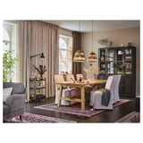 Rustieke eetkamer met houten tafel, grijze stoelen, zwarte kast en houten hangers. Roze gordijnen, kleurrijk tapijt en planten voegen warmte toe.