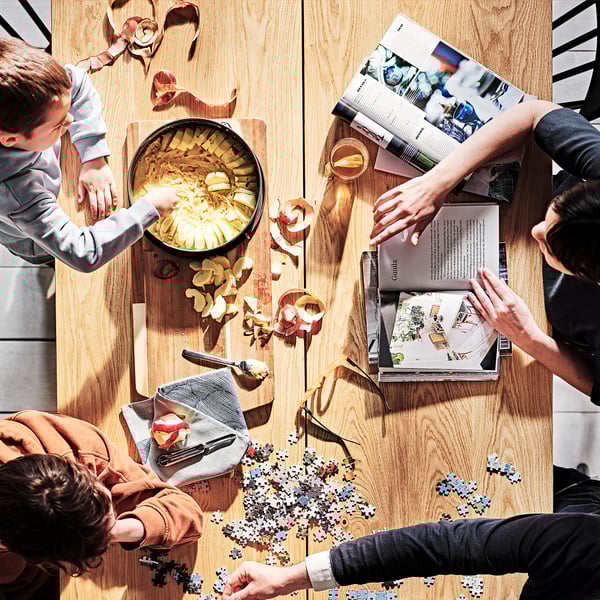 Drie mensen rond MÖCKELBY eiken tafel: koken, lezen, puzzelen.