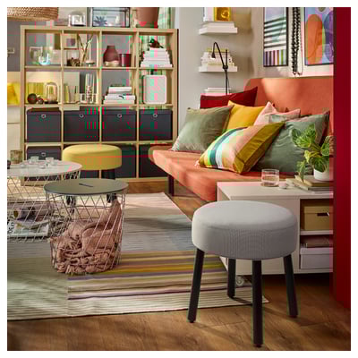Kleurrijke woonkamer: grijze poef, oranje bank, gele kruk, veelkleurig vloerkleed, houten planken met decor.