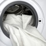 Afbeelding toont een witte matrasbeschermer met een gewatteerd patroon in een droger. Het is ontworpen om een steviger slaapoppervlak te bieden en de matras te beschermen.