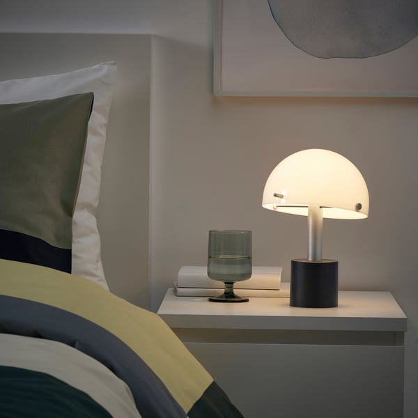 Moderne slaapkamer met een NÖDMAST lamp op het nachtkastje; De lamp is klein, wit en koepelvormig, naast een groen glas en een boek.
