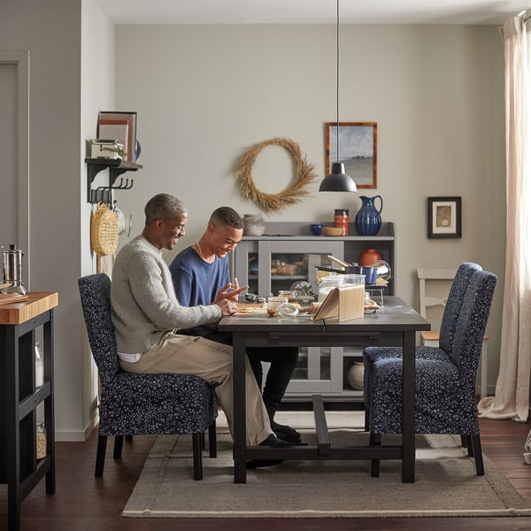Twee mannen zitten aan een zwarte NORDVIKEN-eettafel met blauw gespikkelde stoelen en genieten van een maaltijd in een gezellige kamer met lichtbeige muren en inrichting.