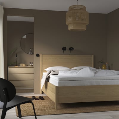 Minimalistische slaapkamer: licht houten bed, wit linnengoed, geweven hanglamp, donkere muren, zwarte stoel, beige vloerkleed.