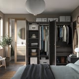 Moderne slaapkamer met zwarte PAX kledingkast, hangende kleding, planken en een grote spiegel. Grijs bed en lichtpunt.