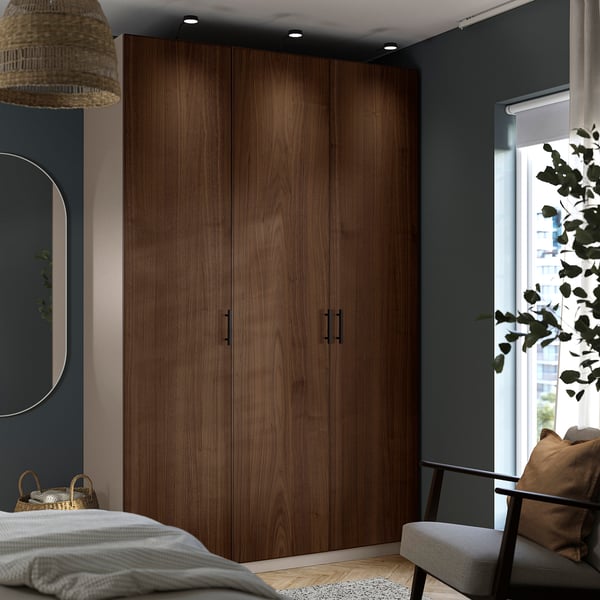 Grote houten kast domineert de hoek. In de buurt van bed, spiegel en raam. Lang, met donkere handvatten. Omgeven door kleinere meubels.