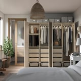 Moderne inloopkast met gespiegelde schuifdeuren, georganiseerde, beige lades, hangende kleding, open garderobe met opbergboxen en een grote plant.