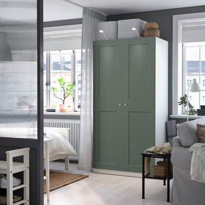Deze afbeelding is een groene PAX-kledingkast in een moderne kamer. Het staat hoog, naast een bank en een rek, en biedt voldoende opbergruimte met zijn twee deuren.
