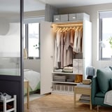 Open kledingkast met hangers, opgevouwen kleding en planken met opbergdozen in een goed verlichte kamer met een raam en een groene stoel.
